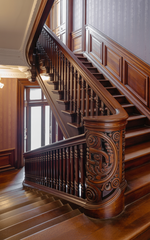 An elegant and natural-looking realistic photograph of a grand staircase featuring an ornate carved mahogany wood banister with intricate scrollwork and slim wooden spindles, polished dark wood treads and a softly patterned hallway backdrop decorated in Victorian revival style with natural lighting