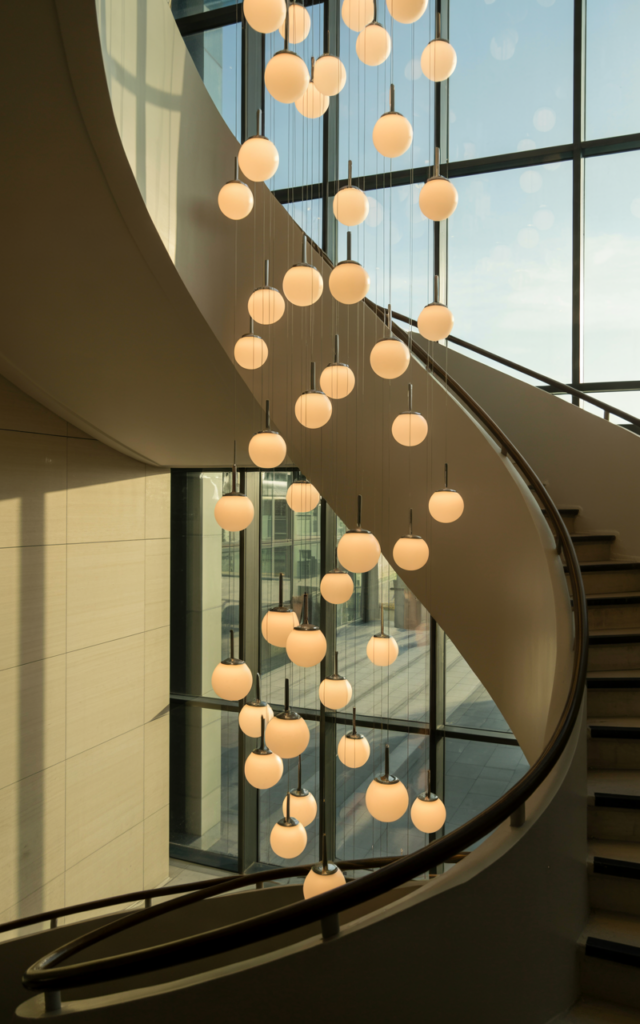 An elegant and natural-looking realistic photograph of a tall stairwell featuring a cascading chandelier with multiple glass globe lights hanging at different heights beside a sweeping staircase, creating a floating sculpture effect above the steps, decorated in modern luxury interior style with natural lighting